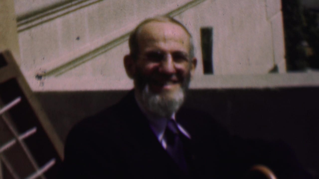In a 1961 San Diego home-movie frame, a smiling bearded man in a dark suit and tie stands near a stone building with a partial grid structure at the edge.