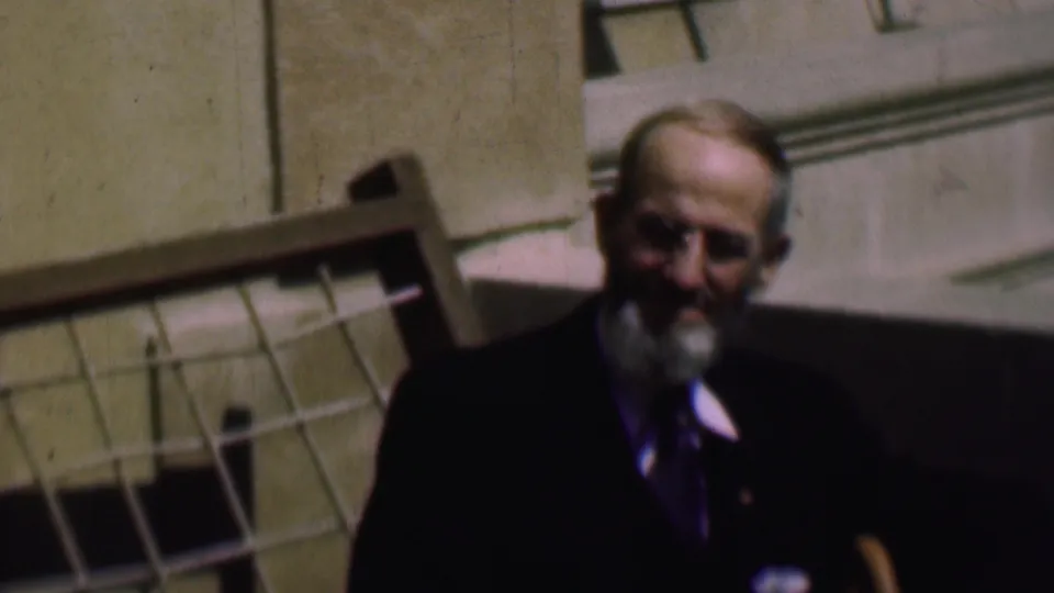 A softly focused 1961 frame shows the bearded man in formal clothing smiling toward the camera beside the stone exterior and grid-like feature.
