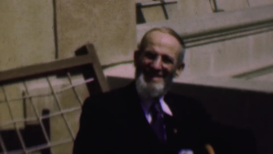 The final frame holds on the smiling bearded man in a dark suit and tie, with the stone building and grid-patterned structure still visible behind him.
