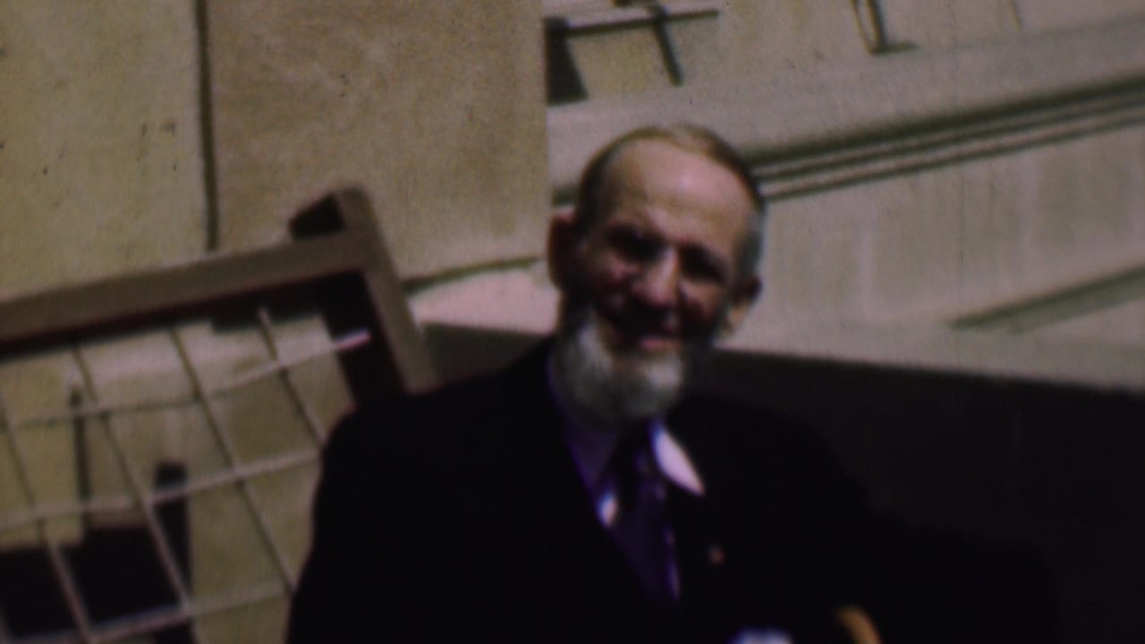 In this 1961 San Diego home-movie frame, a bearded man in glasses and a dark suit faces the camera while seated indoors beside a wooden chair back.