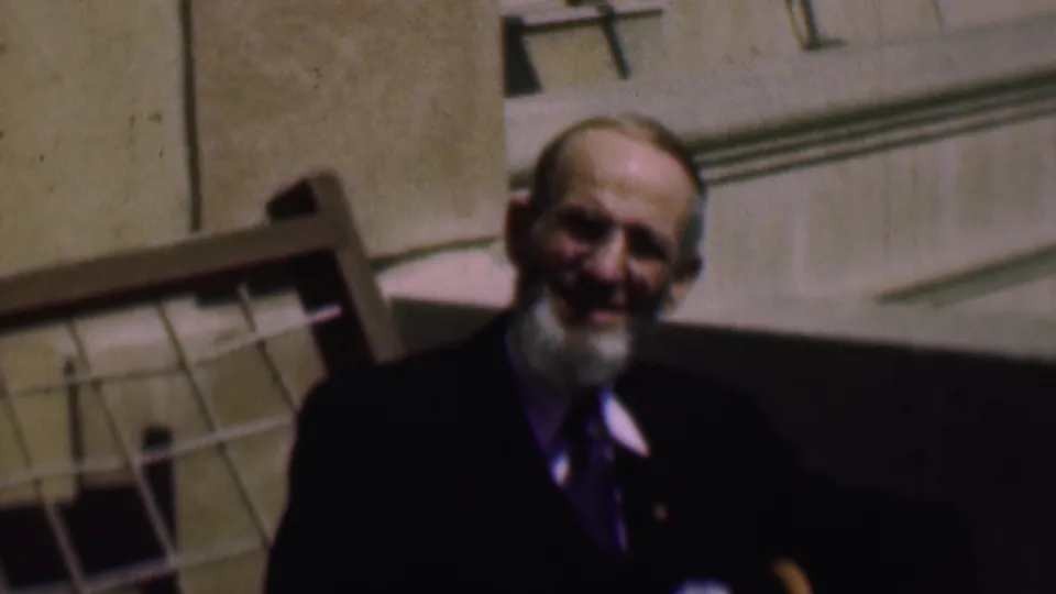 In this 1961 San Diego home-movie frame, a bearded man in glasses and a dark suit faces the camera while seated indoors beside a wooden chair back.
