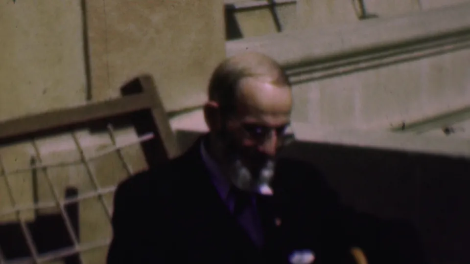 The frame shows the suited, bearded man in glasses leaning or looking downward in the indoor 1961 San Diego home-movie footage.