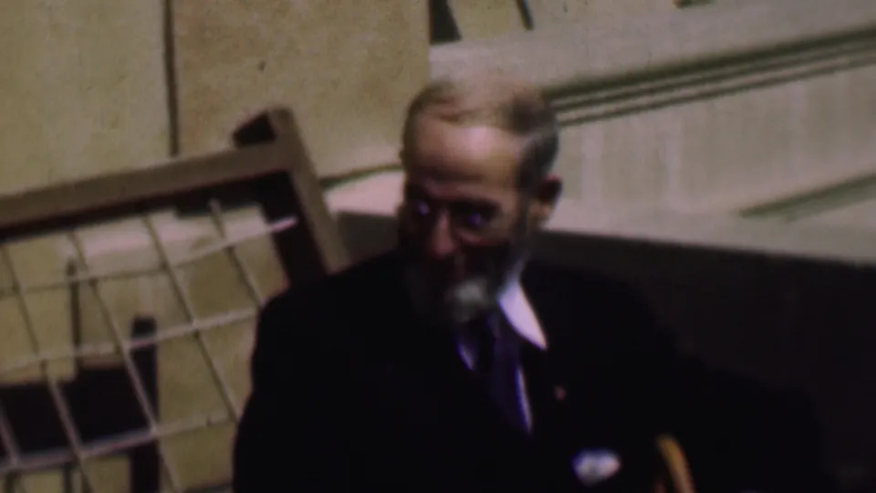 At the end of the clip, the bearded man with glasses sits partly in shadow near a wooden chair back in the 1961 indoor home-movie scene.