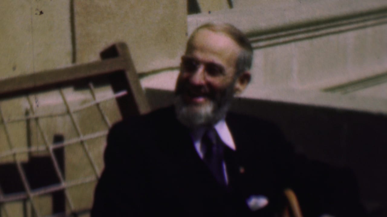 In 1961 San Diego home-movie footage, an older white-bearded man in a dark suit and tie sits outdoors beside a pale stone building and a wooden chair or railing.