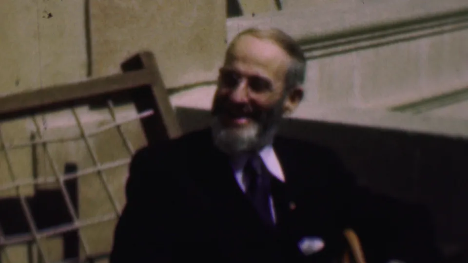 In 1961 San Diego home-movie footage, an older white-bearded man in a dark suit and tie sits outdoors beside a pale stone building and a wooden chair or railing.