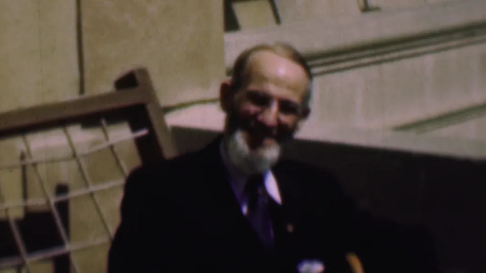 The older man smiles toward the home-movie camera, wearing a dark suit, white shirt, and tie against a sunlit building exterior in the supplied 1961 San Diego clip.
