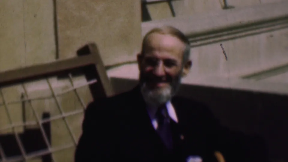 A softly focused 8mm frame shows the older bearded man seated outdoors in formal clothing, with a chair or railing and pale building facade behind him.