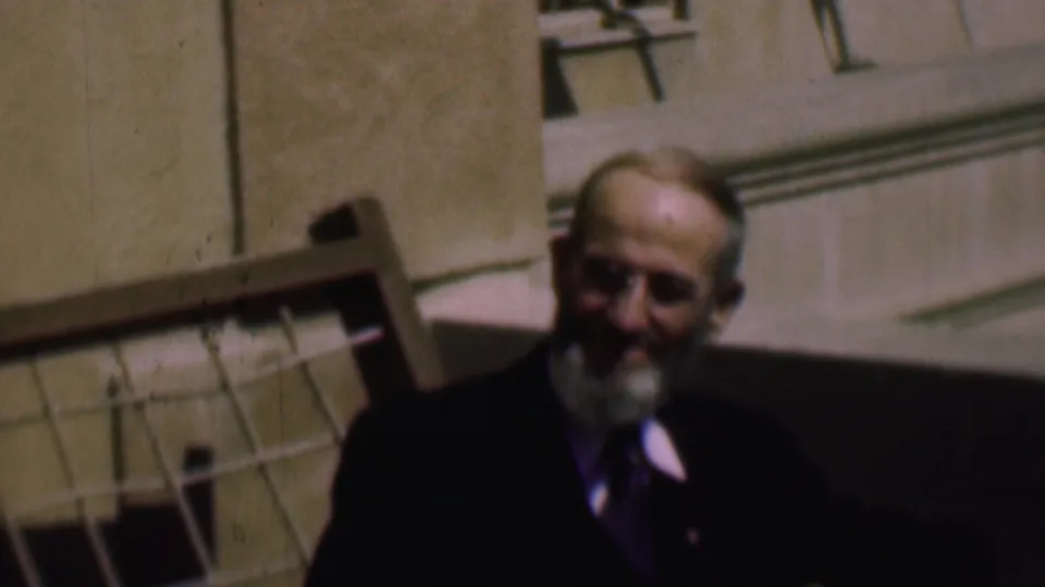 The final frame holds on the older man seated outdoors, his face partly in shadow, with the wooden chair or railing and light-colored building still visible behind him.