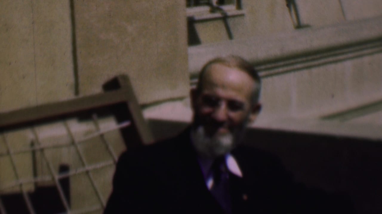 In supplied 1961 San Diego home-movie footage, two older bearded men appear outside a building, one in a dark suit and tie and one partly shadowed near a leaning chair.