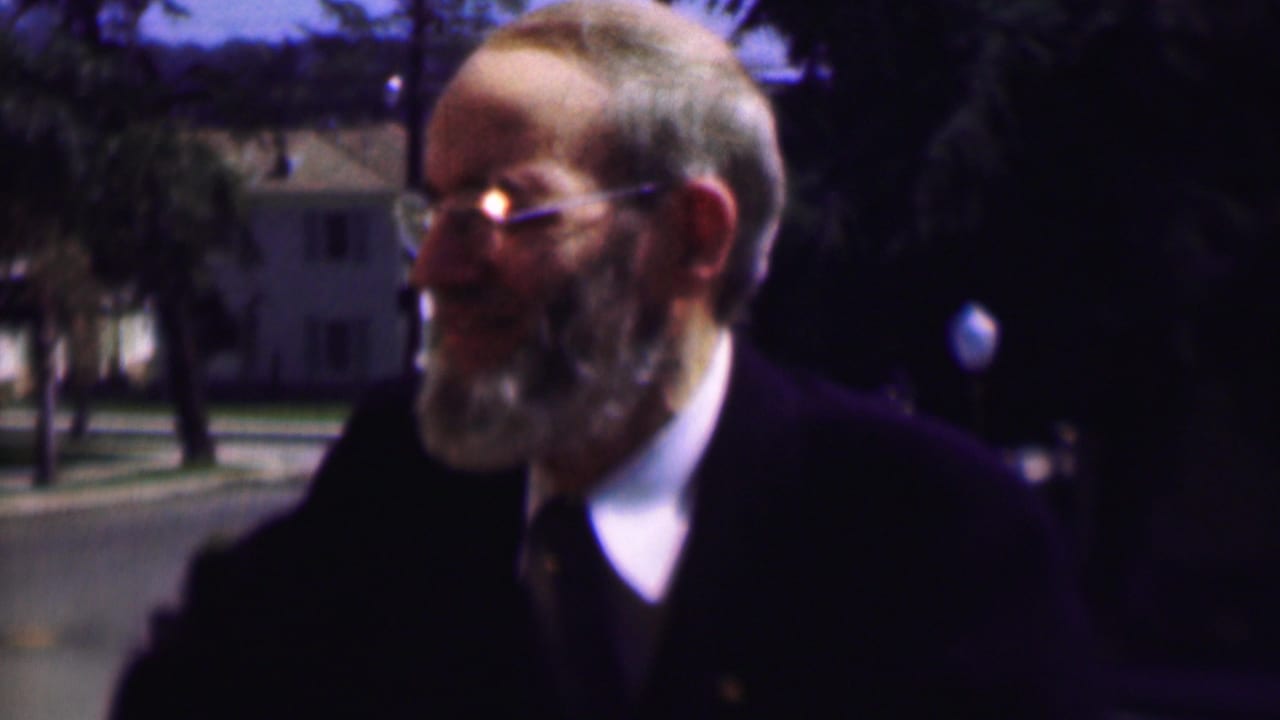Older bearded man in glasses and a dark suit smiles outdoors near trees and houses.