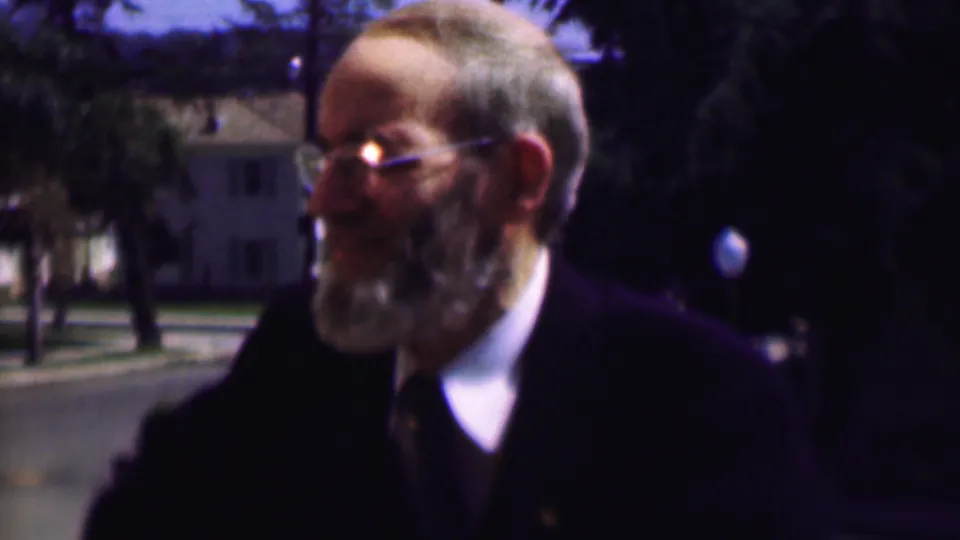 In the supplied 1961 San Diego footage, an older man with a gray beard and glasses wears a dark suit and tie while smiling outdoors near trees, a street, and distant houses.