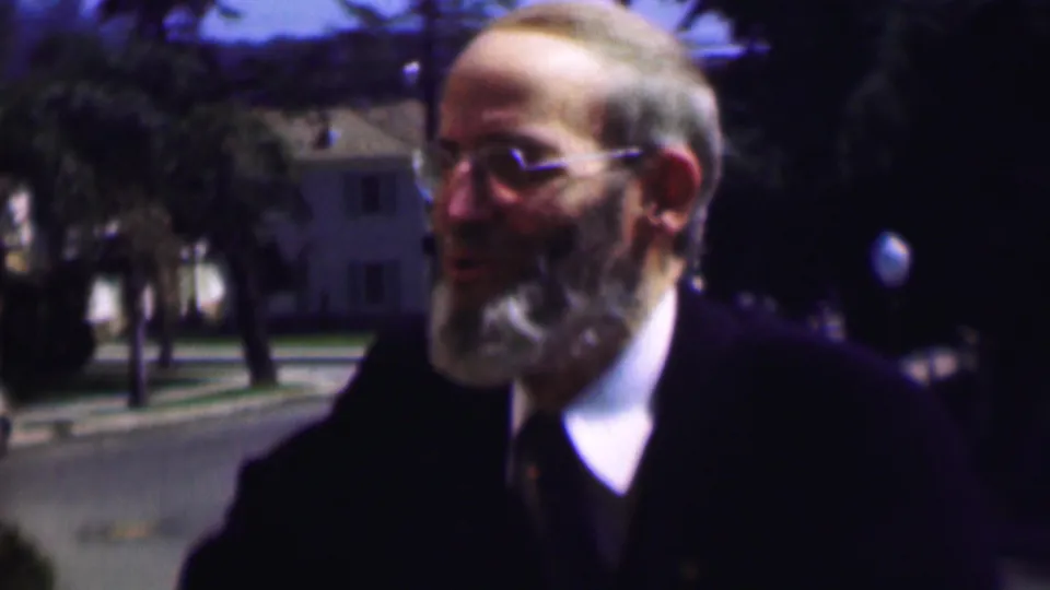The older bearded man in glasses is seen in partial profile in the supplied 1961 San Diego home movie, with trees, a roadway, and houses behind him.