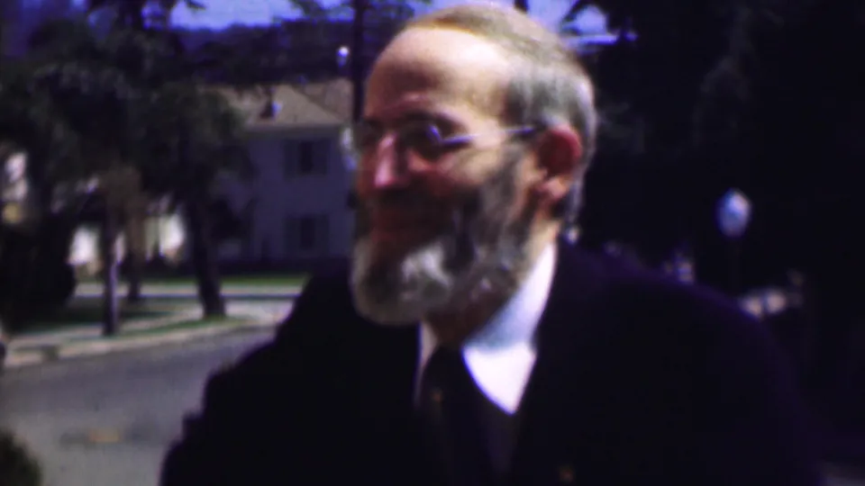 The older gentleman with glasses and a gray beard smiles in the supplied 1961 San Diego footage, framed close to the camera with a tree-lined street and houses in the background.