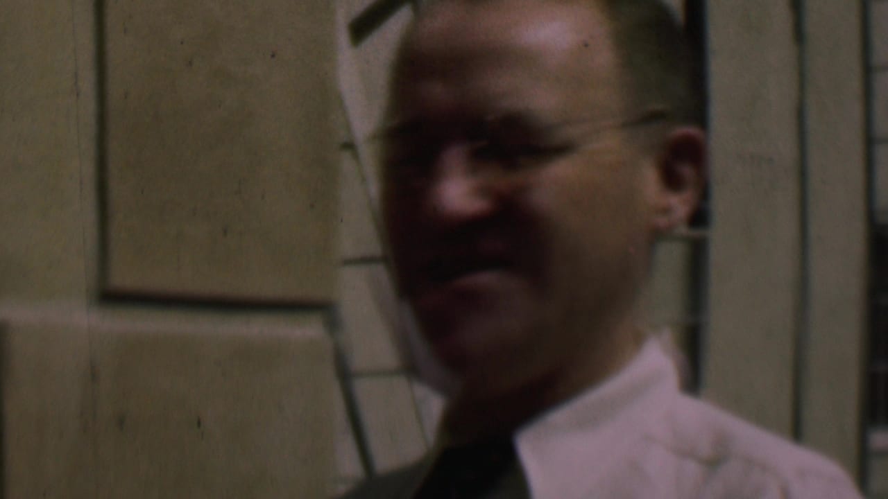 Adult man in glasses, white shirt, and dark tie standing outside a cream-colored building in 1961 San Diego footage.