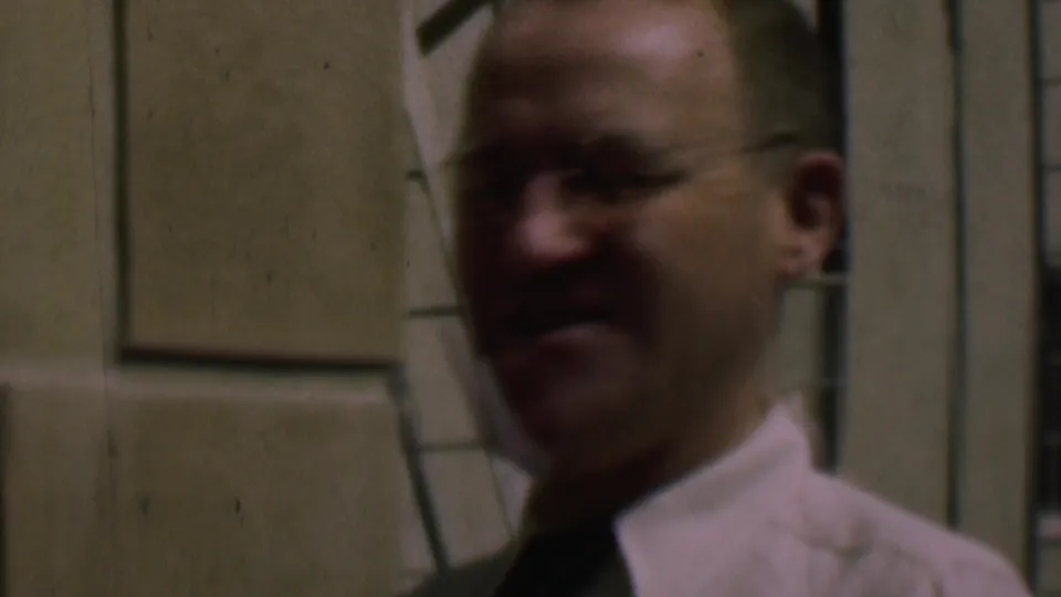Adult man in glasses, white shirt, and dark tie standing outside a cream-colored building in 1961 San Diego footage.