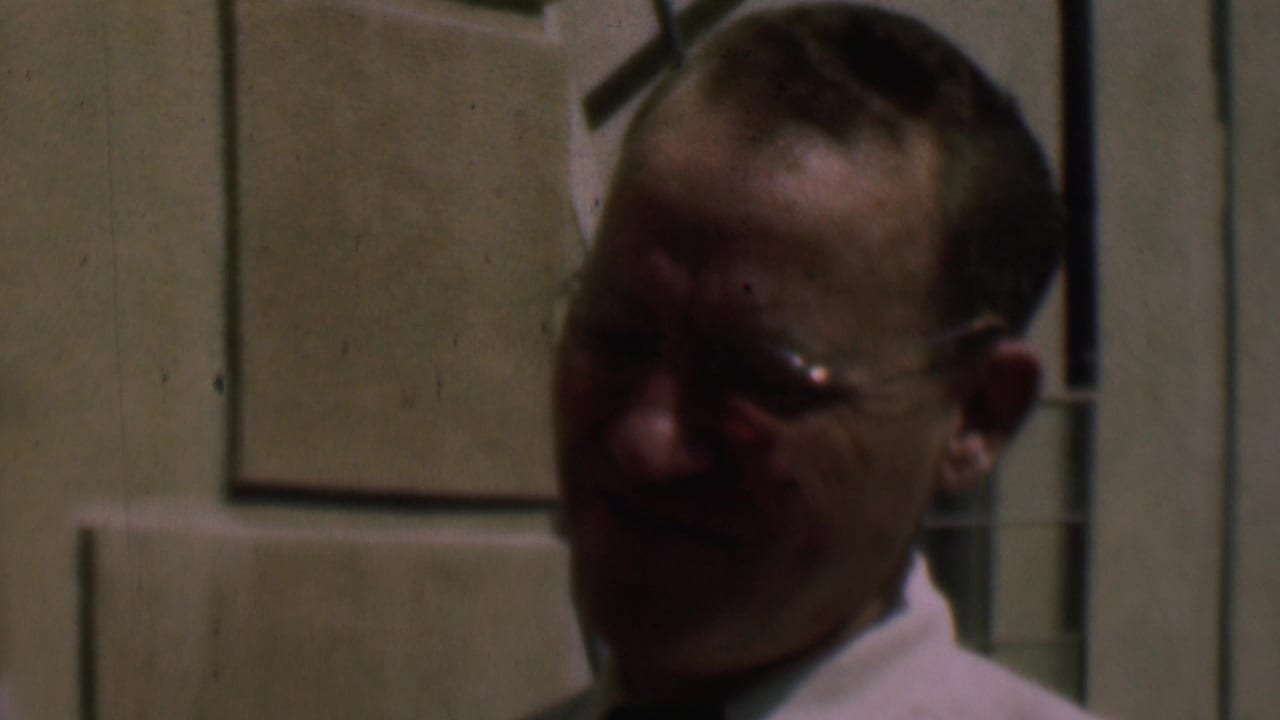 Man in glasses partly in shadow beside a cream-colored wall and window frame.
