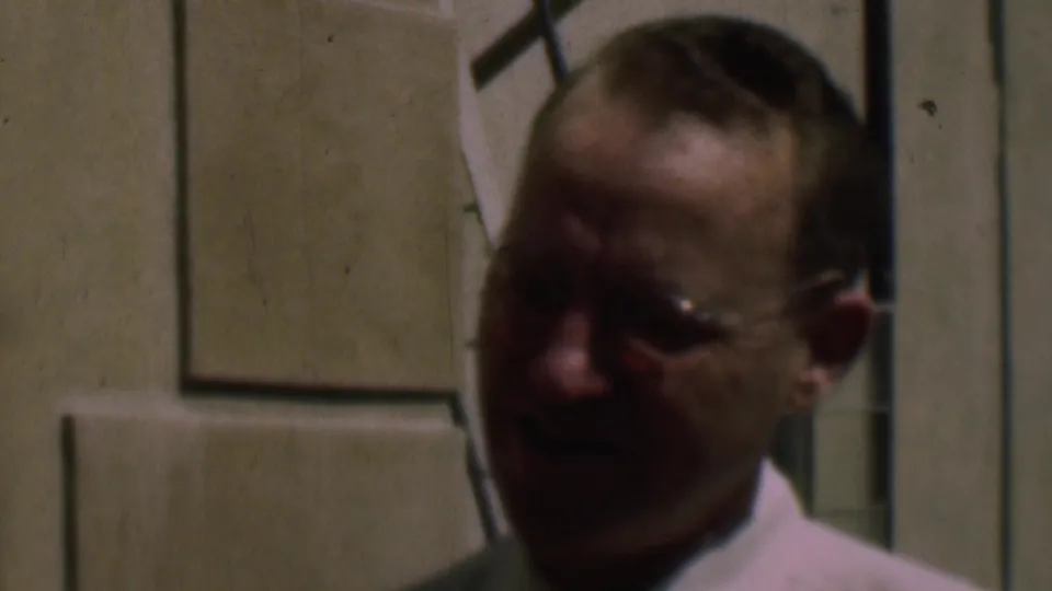 Adult man in a white shirt and glasses seen in soft 8mm close-up outside a cream-colored building in the supplied 1961 San Diego clip.