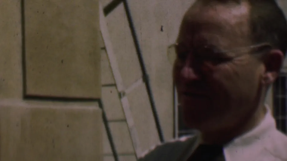 Profile view of the man in glasses and a white shirt near a cream-colored exterior wall and window frame in the 1961 San Diego home-movie footage.