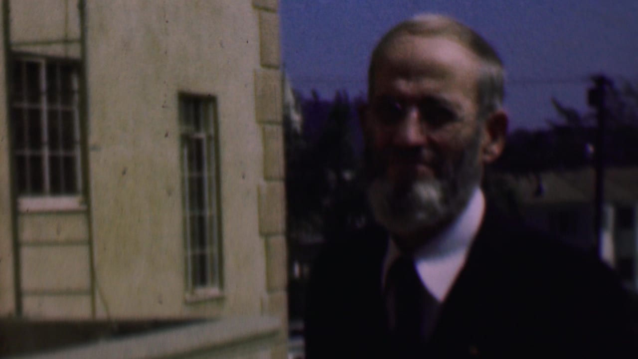 Older bearded man in glasses and a dark jacket stands beside a light-colored building in supported 1961 San Diego home-movie footage.