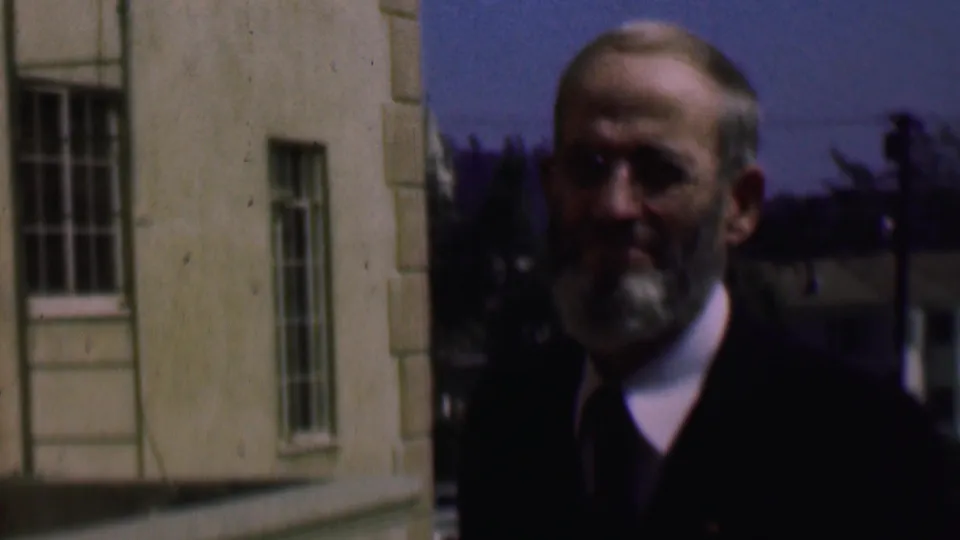 Older bearded man in glasses and a dark jacket stands beside a light-colored building in supported 1961 San Diego home-movie footage.