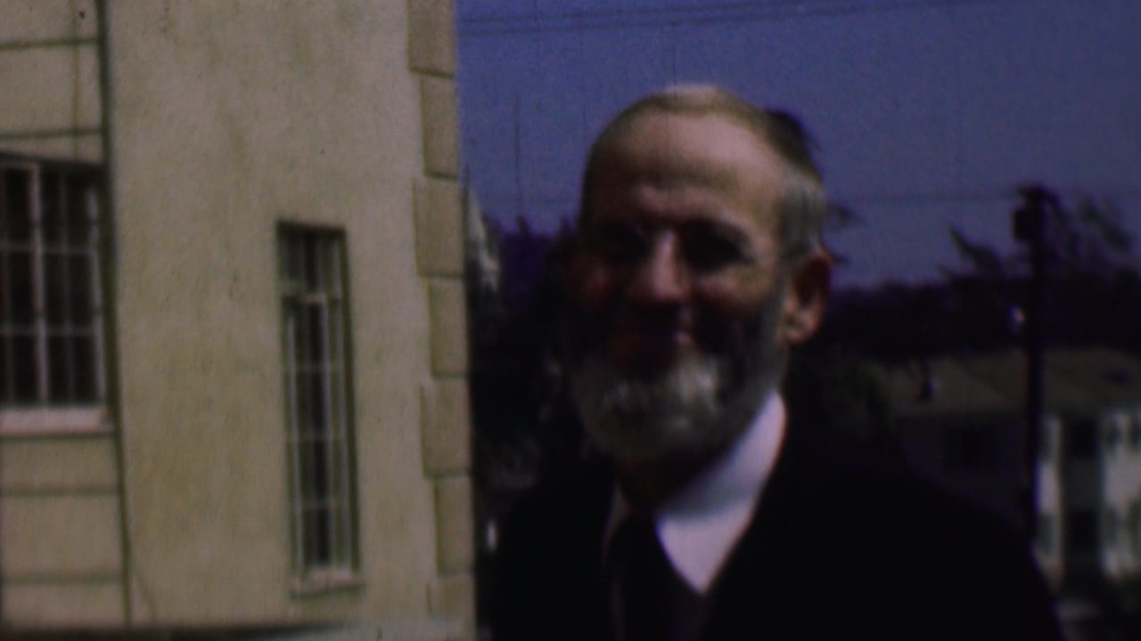 Older man with a white beard faces the camera near a building and trees.