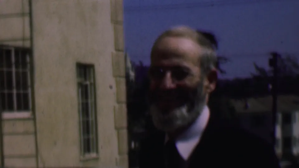 Smiling older man in glasses and a dark jacket stands close to the camera beside barred windows in supported 1961 San Diego home-movie footage.