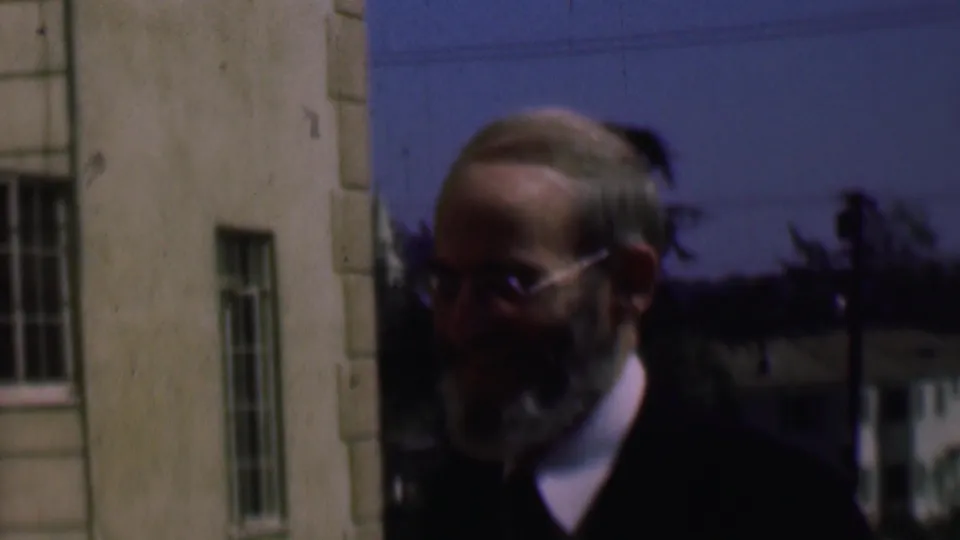 Older bearded man in glasses looks downward near a light-colored building, with trees and houses behind him in 1961 San Diego home-movie footage.
