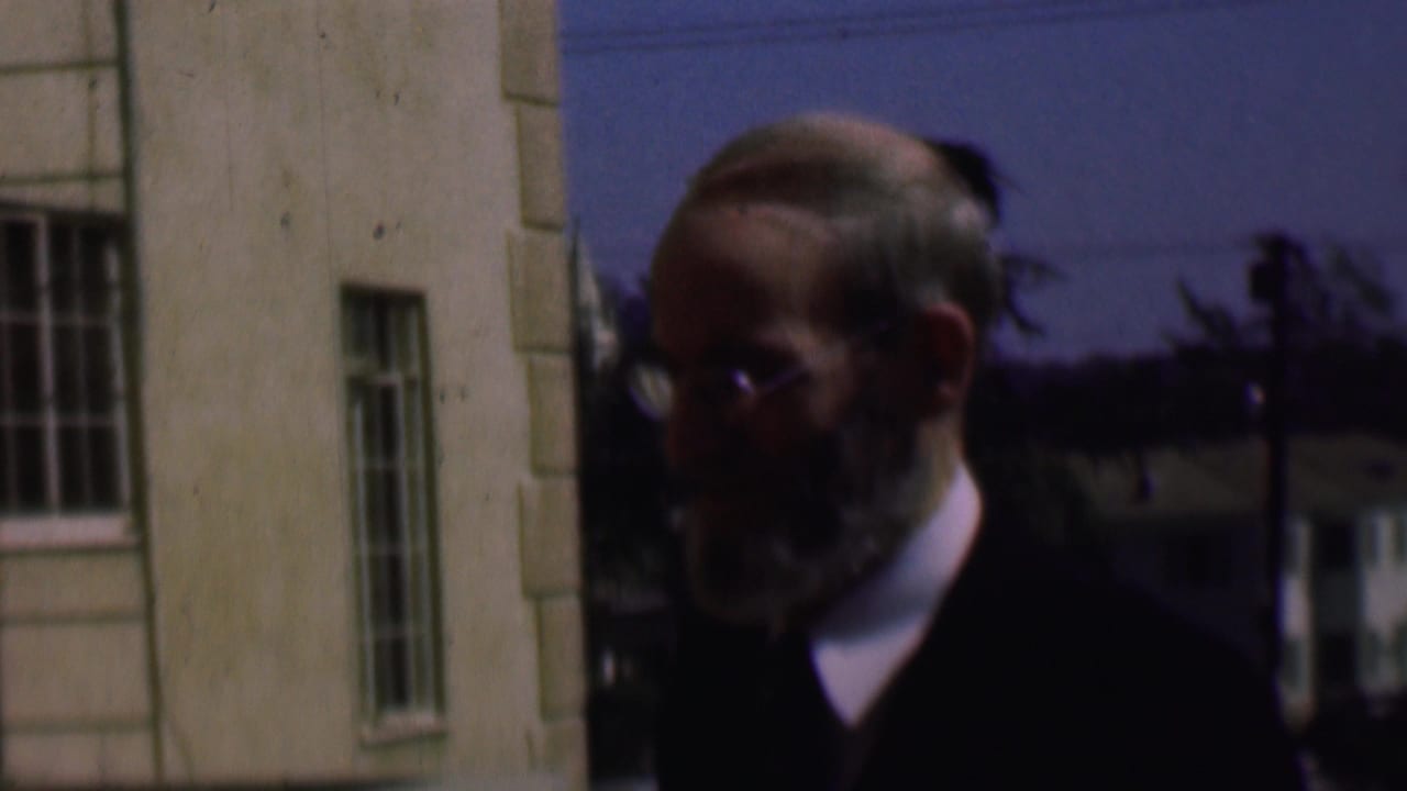 Older bearded man in glasses and a dark jacket stands outdoors near a light-colored building in 1961 San Diego home-movie footage.