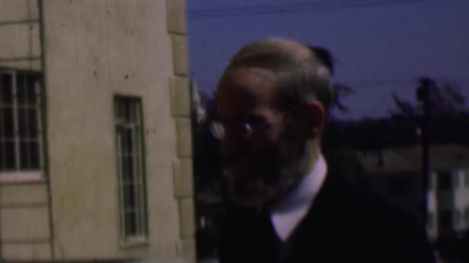 Older bearded man in glasses and a dark jacket stands outdoors near a light-colored building in 1961 San Diego home-movie footage.