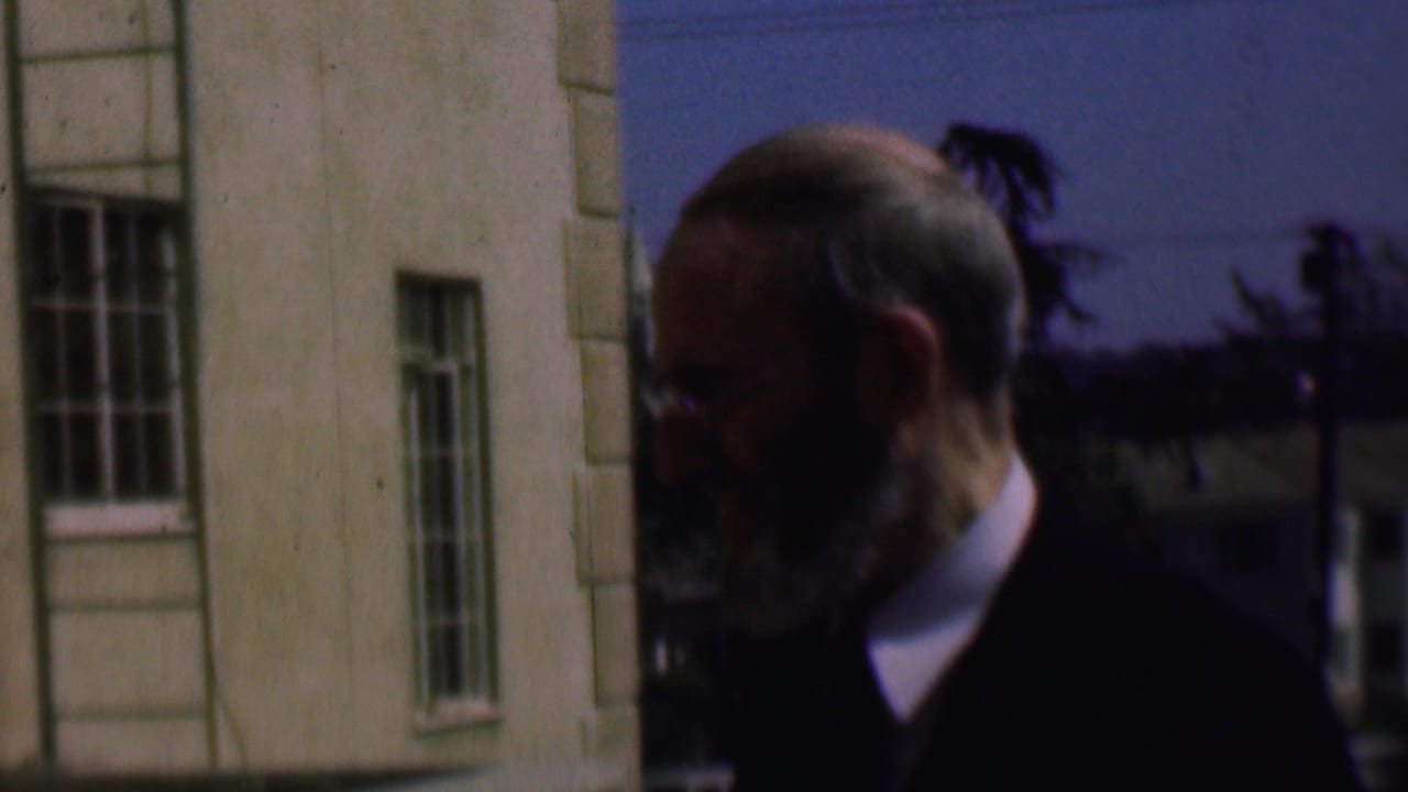 Older man in profile near barred windows and a pale building wall.
