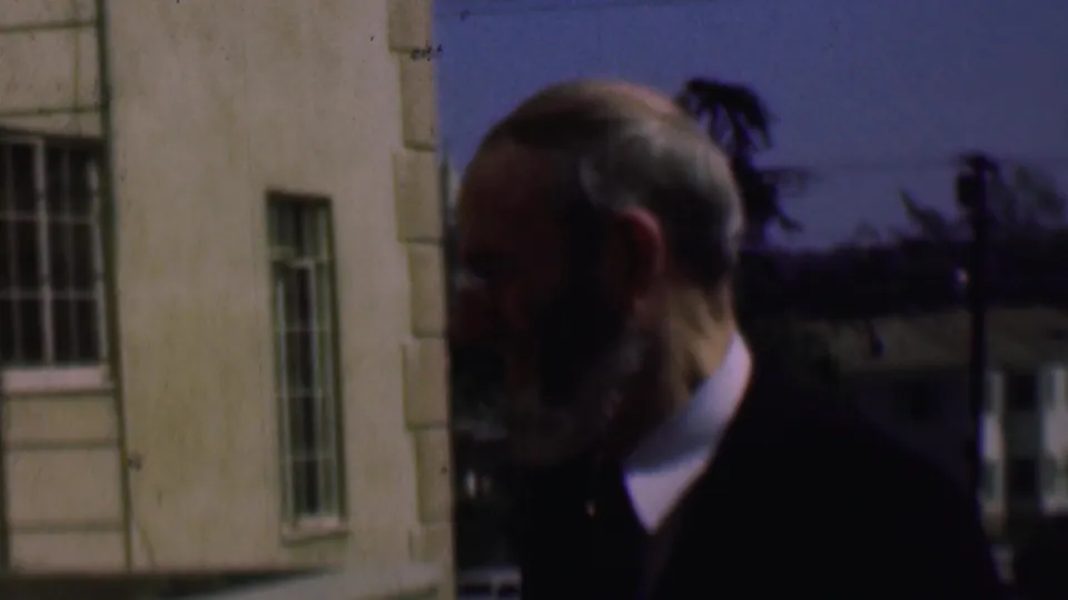 Bearded older man in a dark jacket is filmed close to a light-colored building with narrow barred windows and distant trees.