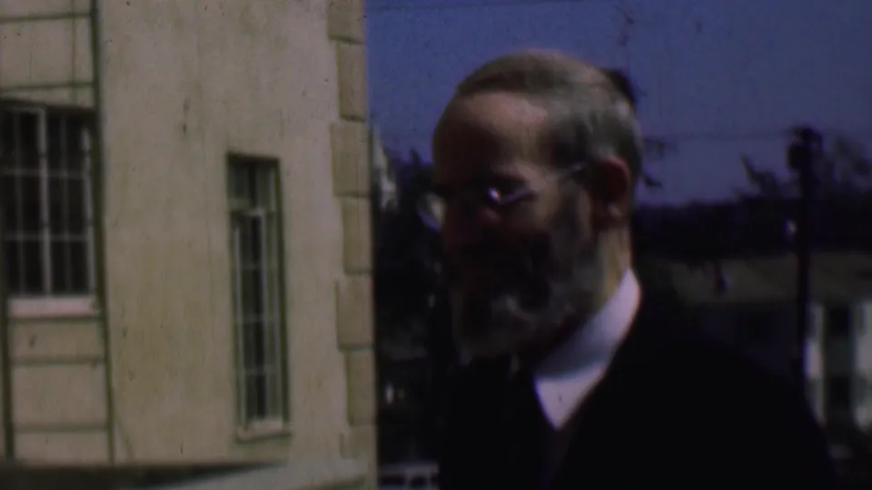 Older bearded man with glasses faces toward the camera outdoors near a pale building, with barred windows and trees in the background.