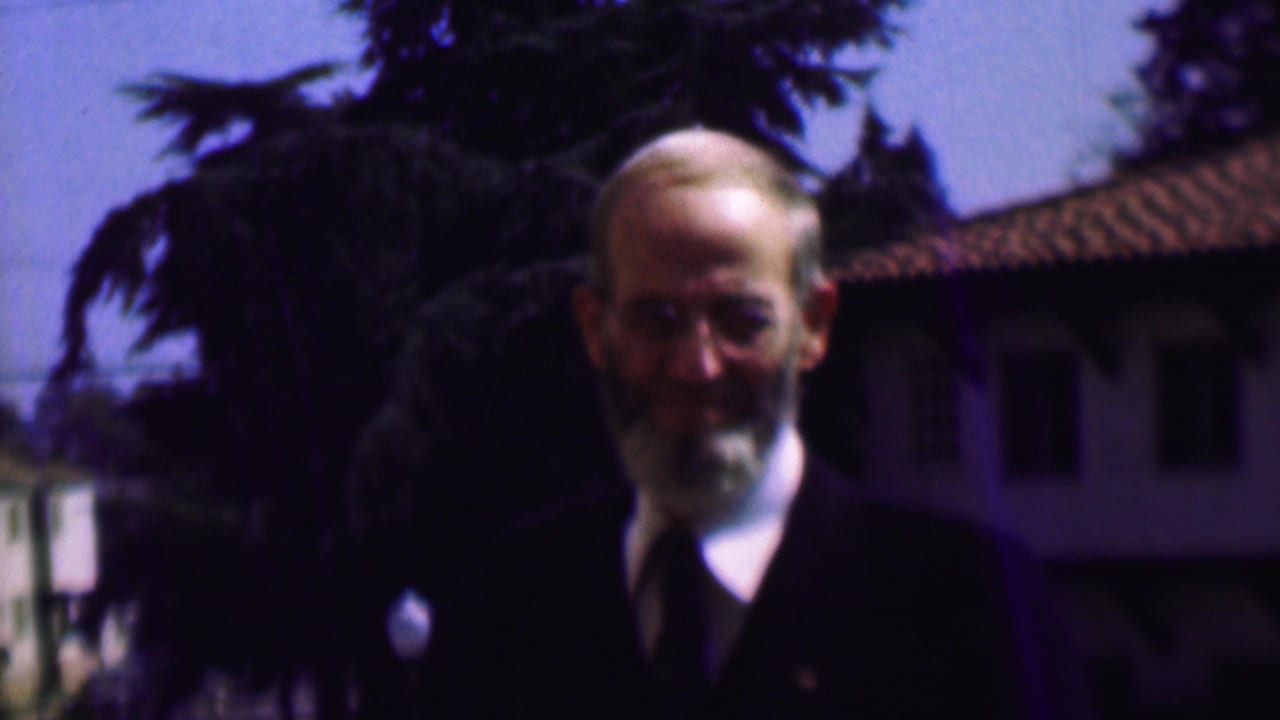In 1961 San Diego home-movie footage, an older bearded man wearing glasses, a dark suit, white shirt, and tie stands outdoors before trees and a tiled-roof building.