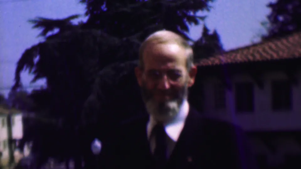In 1961 San Diego home-movie footage, an older bearded man wearing glasses, a dark suit, white shirt, and tie stands outdoors before trees and a tiled-roof building.