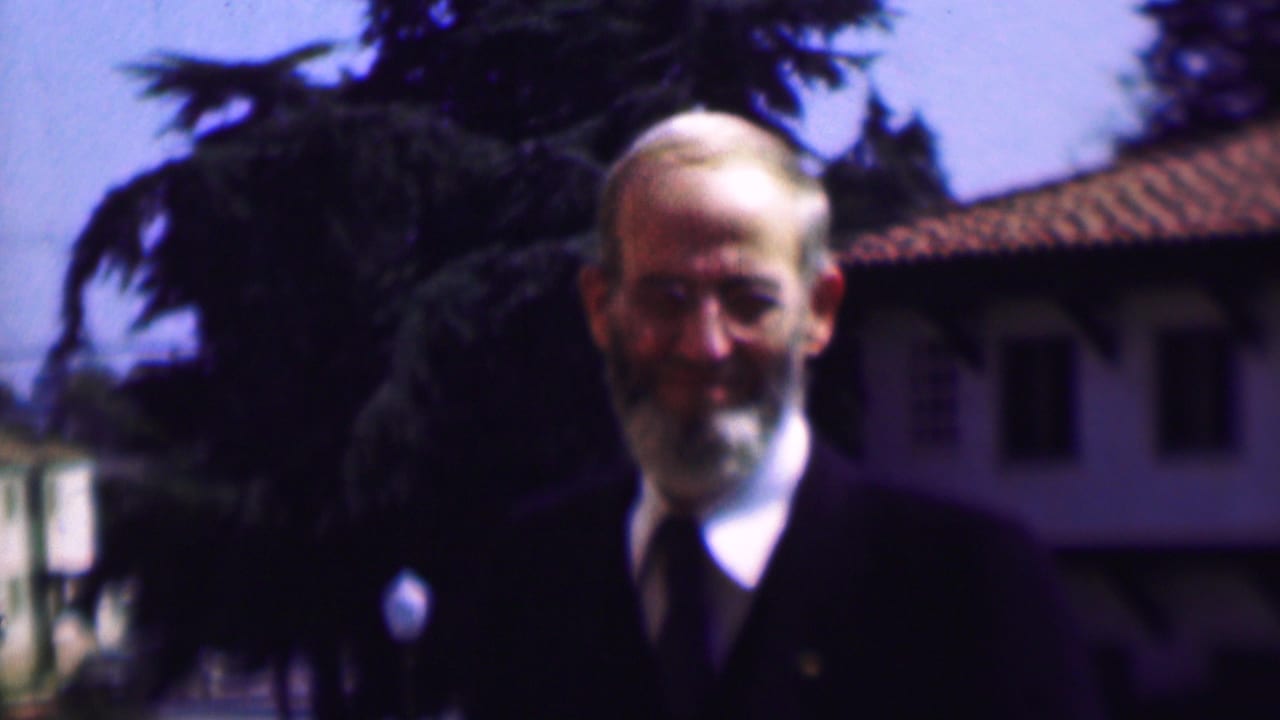 The older man remains framed from the chest up in the 1961 San Diego footage, facing the camera in a dark suit and tie with trees and a tiled roof behind him.