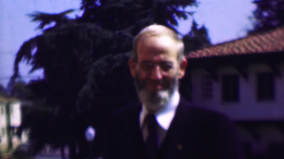 The older man remains framed from the chest up in the 1961 San Diego footage, facing the camera in a dark suit and tie with trees and a tiled roof behind him.