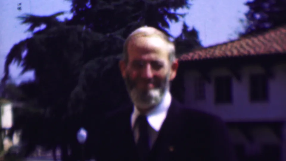 The 1961 San Diego home-movie frame shows the older bearded man smiling slightly while standing outdoors in a dark suit and tie near trees and a stucco-like building facade.