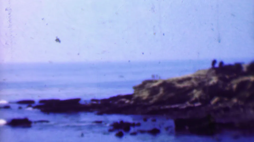 A brief 1961 San Diego coastal view shows rocks, ocean water, a low cliff, and small distant figures silhouetted near the right edge of the frame.