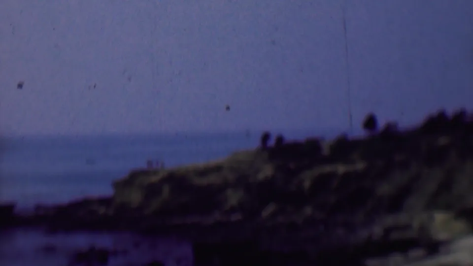 1961 San Diego-area 8mm frame of a rocky ocean bluff under a hazy sky, with small silhouetted people visible near the top edge of the cliff.