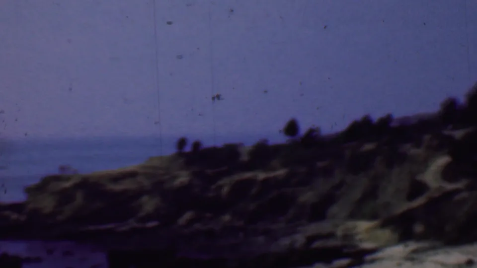 1961 San Diego-area 8mm frame showing a wider view of the rocky bluff, ocean at left, and distant visitors silhouetted against the sky.