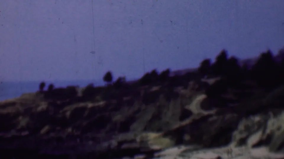 1961 San Diego-area 8mm frame showing a hazy coastal slope with erosion lines or paths, dark shoreline rocks, ocean water, and distant people on the bluff.