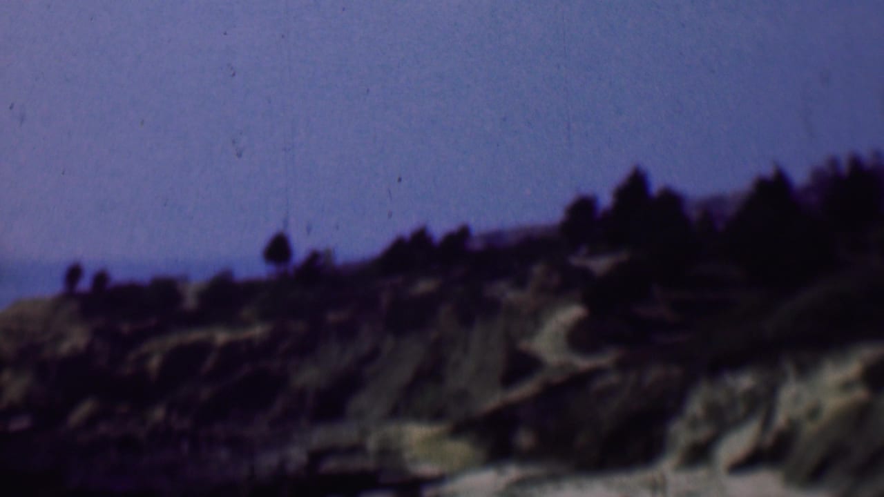 1961 San Diego home-movie frame showing a rugged hillside with pale rocky cuts, sparse vegetation, and a dark tree line silhouetted against blue sky.