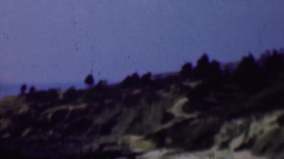 1961 San Diego home-movie frame showing a rugged hillside with pale rocky cuts, sparse vegetation, and a dark tree line silhouetted against blue sky.
