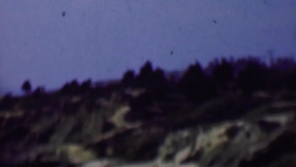 1961 San Diego 8mm frame continuing the pan across a scrubby hillside, with dark ridge trees and pale eroded ground below a blue sky.