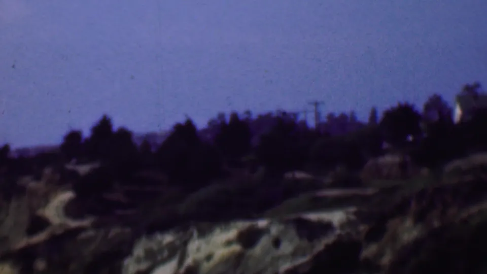 1961 San Diego home-movie frame showing a wider hillside view with dark trees, pale exposed terrain, and faint utility poles near the ridge.