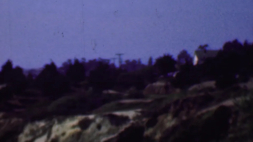 Final 1961 San Diego frame of the pan showing a darkened hillside ridge, scattered trees, brush, exposed pale soil, and faint utility poles against the sky.