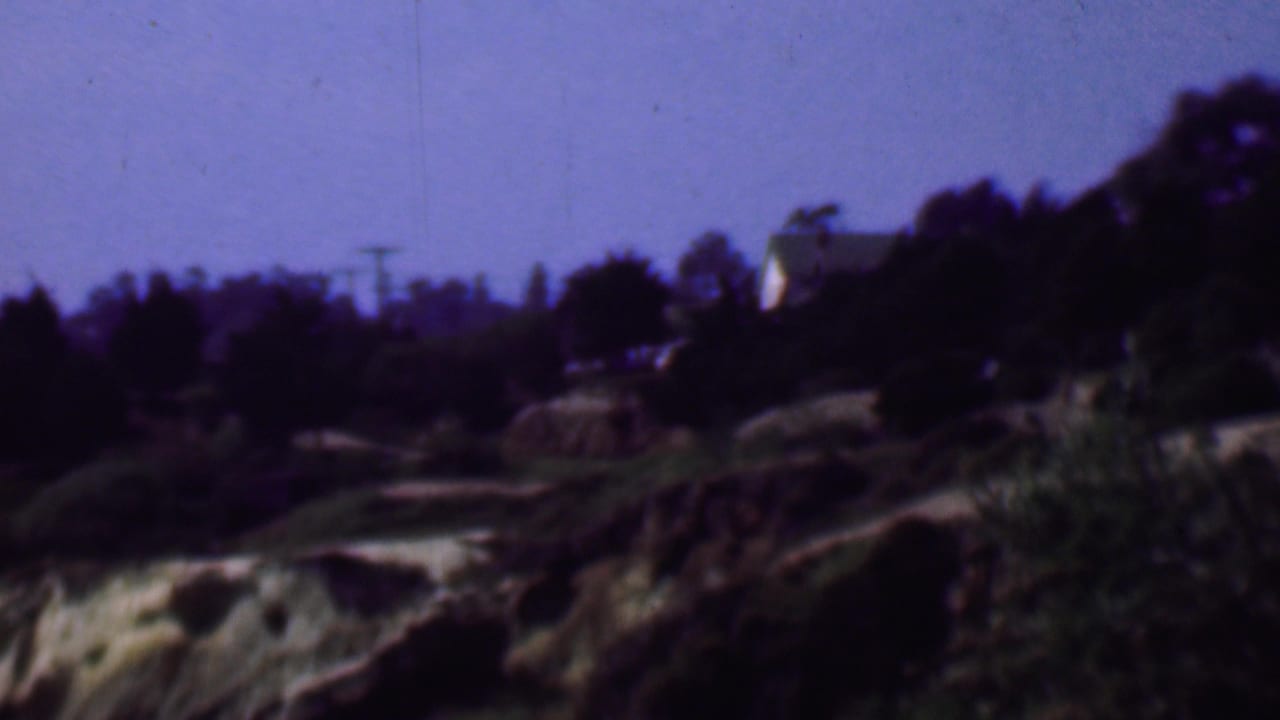 Soft 1961 San Diego home-movie frame showing a distant light-colored hillside house partly obscured by trees, with rocky slopes and dark vegetation in the foreground.