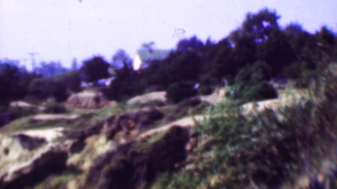 1961 San Diego-area 8mm frame showing rocky coastal terrain with green brush, tall trees, hazy purple sky, utility lines, and a distant pale building.