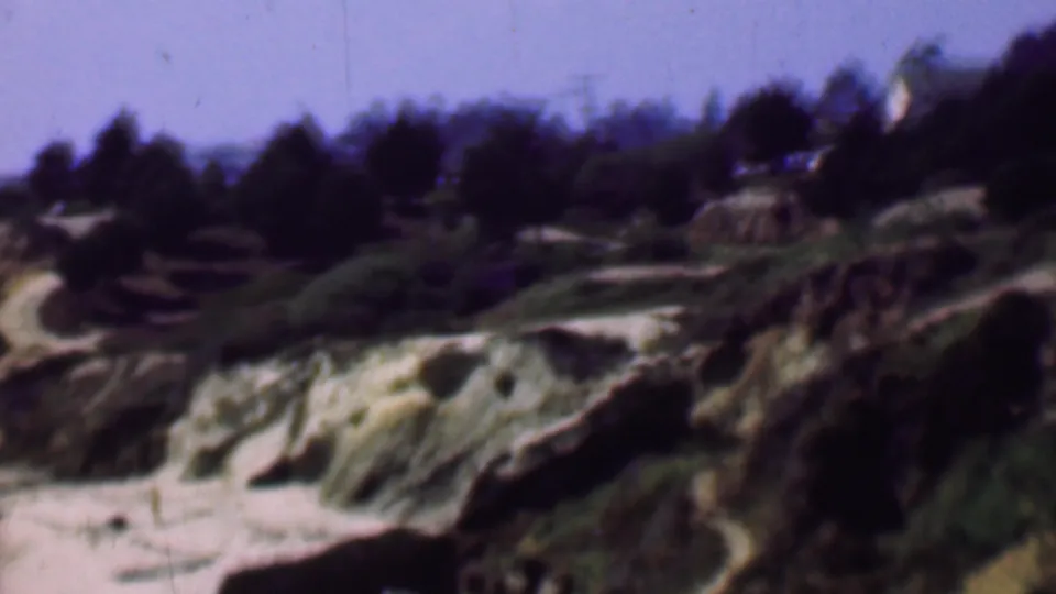 Wide 1961 San Diego-area home-movie frame of coastal bluffs with sandy tracks, rocky outcrops, shrubs, a tree-covered horizon, and soft hazy sky.