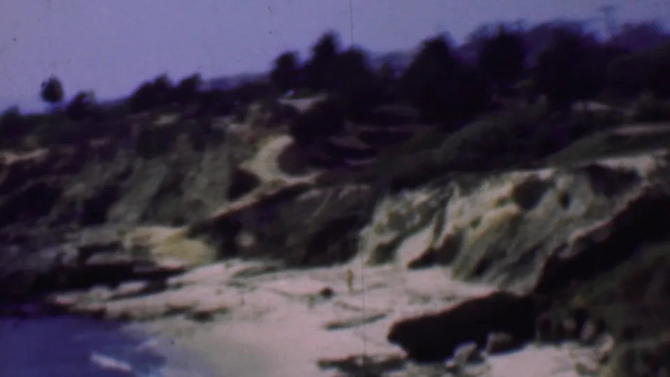 1961 San Diego-area 8mm frame showing a rocky shoreline with small waves, sandy beach, eroded coastal slopes, brush, trees, and hazy sky.